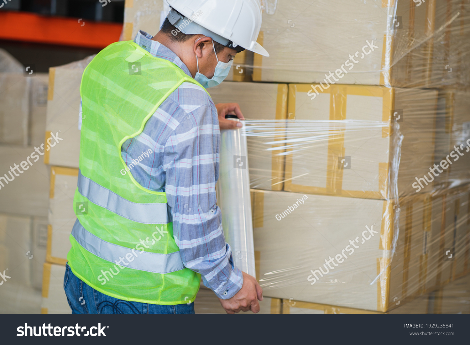 close up view of man worker wearing protective face mask and safety suite wrapping stretch film parcel on pallet in factory warehouse, logistic industry concept.  #1929235841