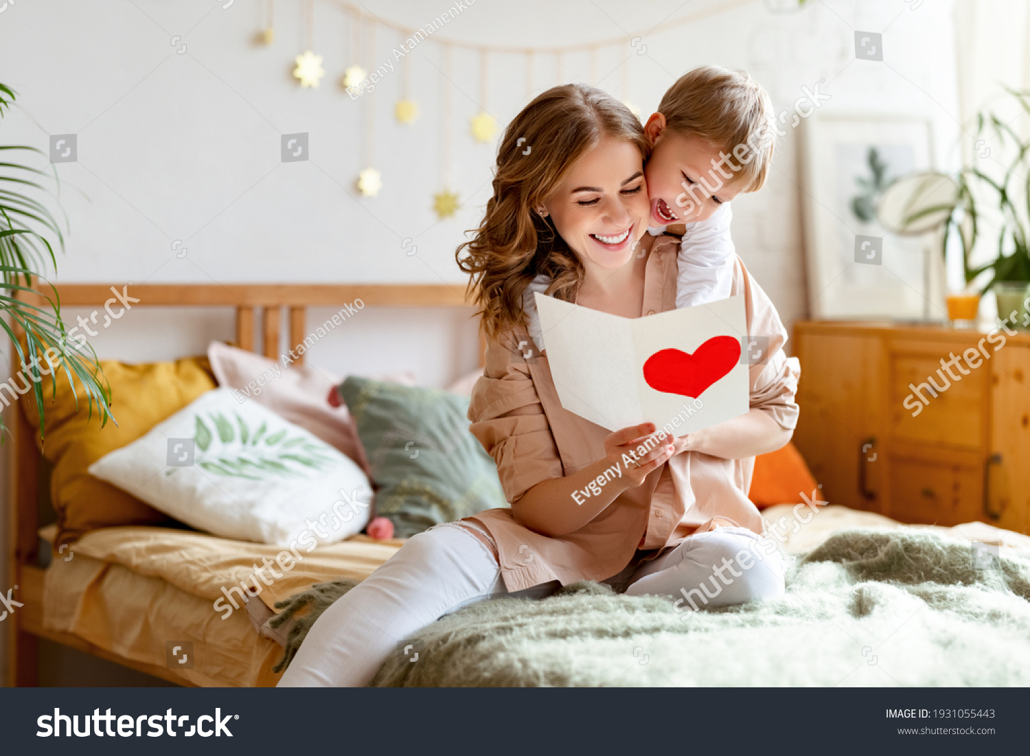 Cheerful mother hugging son and reading handmade greeting card with heart while resting on bed during holiday celebration mothers day  at home #1931055443