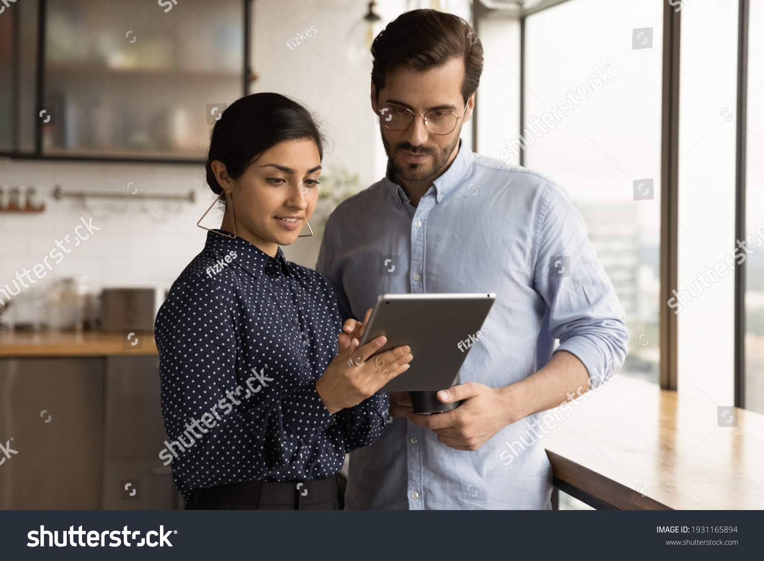 Close up diverse colleagues using tablet together, discussing online project, smiling Indian businesswoman and Caucasian businessman wearing glasses looking at device screen, standing in office #1931165894