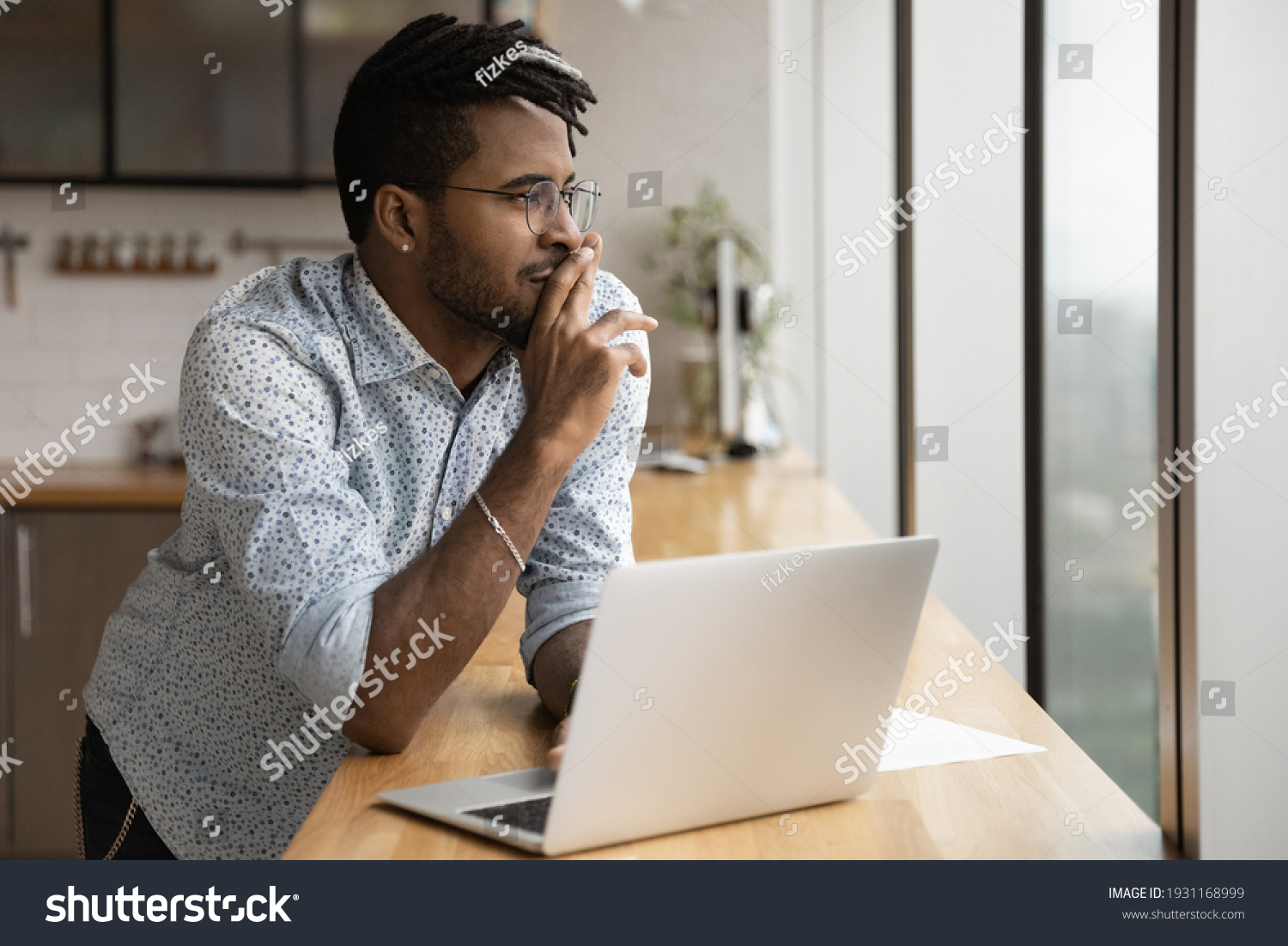 Close up dreamy African American man wearing glasses looking to aside out panoramic window, visualizing, pondering new opportunities or online project strategy, standing at table with laptop #1931168999