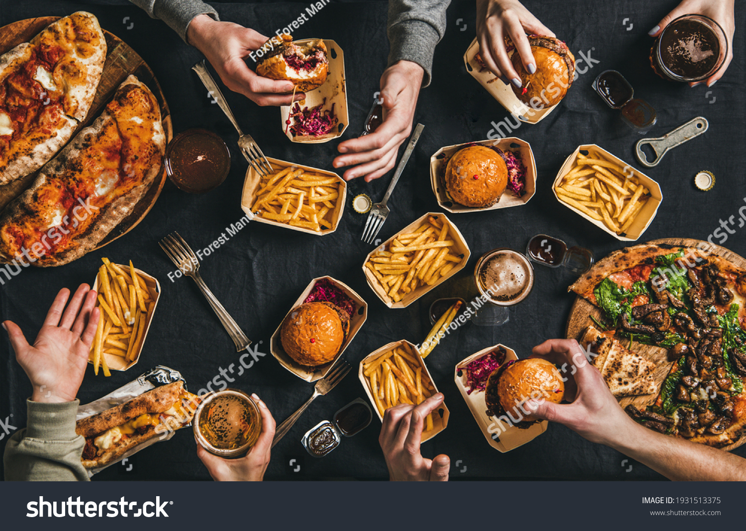 Lockdown fast food dinner from delivery service. Flat-lay of peoples hands eating burger, fries, sandwiche, pizza, drinking beer over table background, top view. Quarantine home party, takeaway food #1931513375