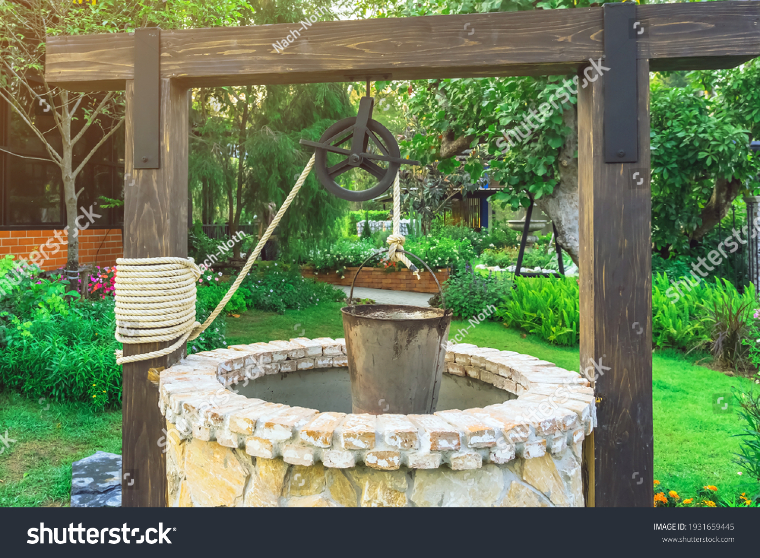 Beautiful artesian well made by stones and wheel pulley with metal bucket and rope in peaceful garden atmosphere. Retro stone water well in rural area. Garden decoration with antique items. #1931659445