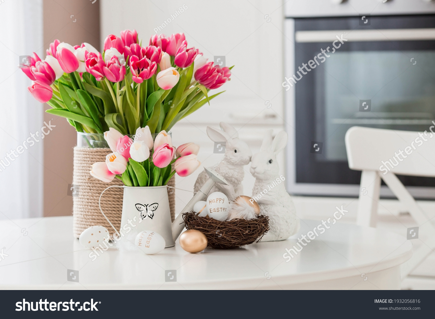 A bouquet of tulips, Easter bunnies and eggs with a golden pattern on the table. In the background is a white Scandinavian-style kitchen. Beautiful greeting card. The minimal concept. #1932056816