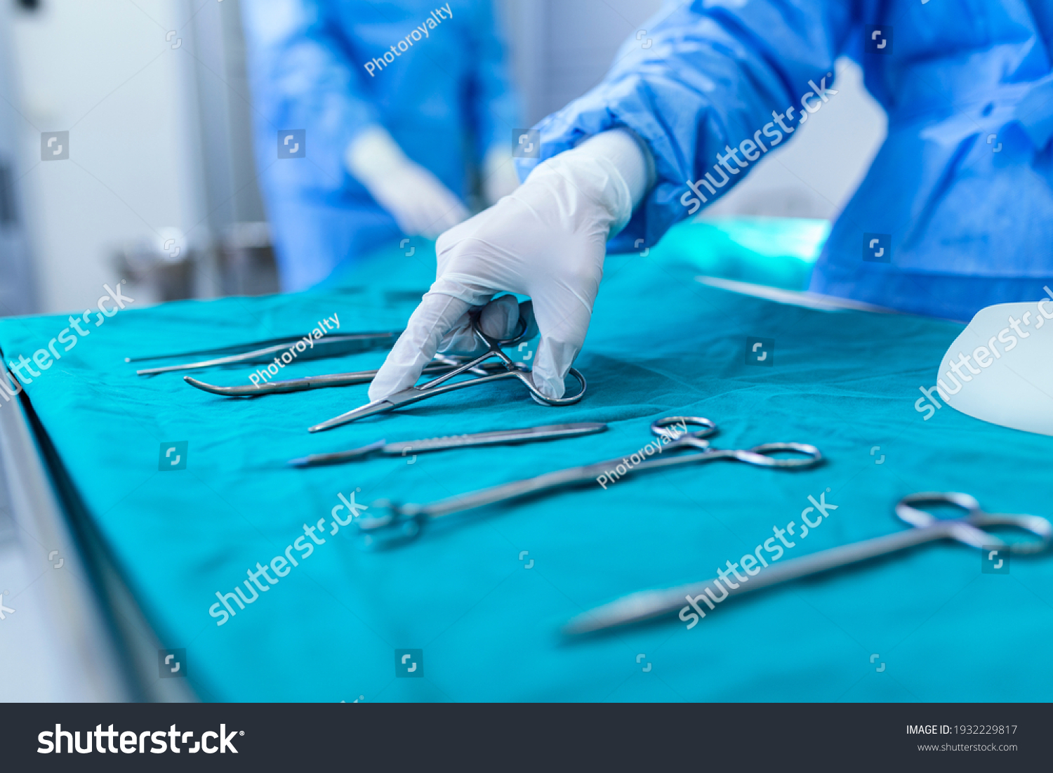 Nurse hand taking surgical instrument for group of surgeons at background operating patient in surgical theatre. Steel medical instruments ready to be used. Surgery and emergency concept #1932229817