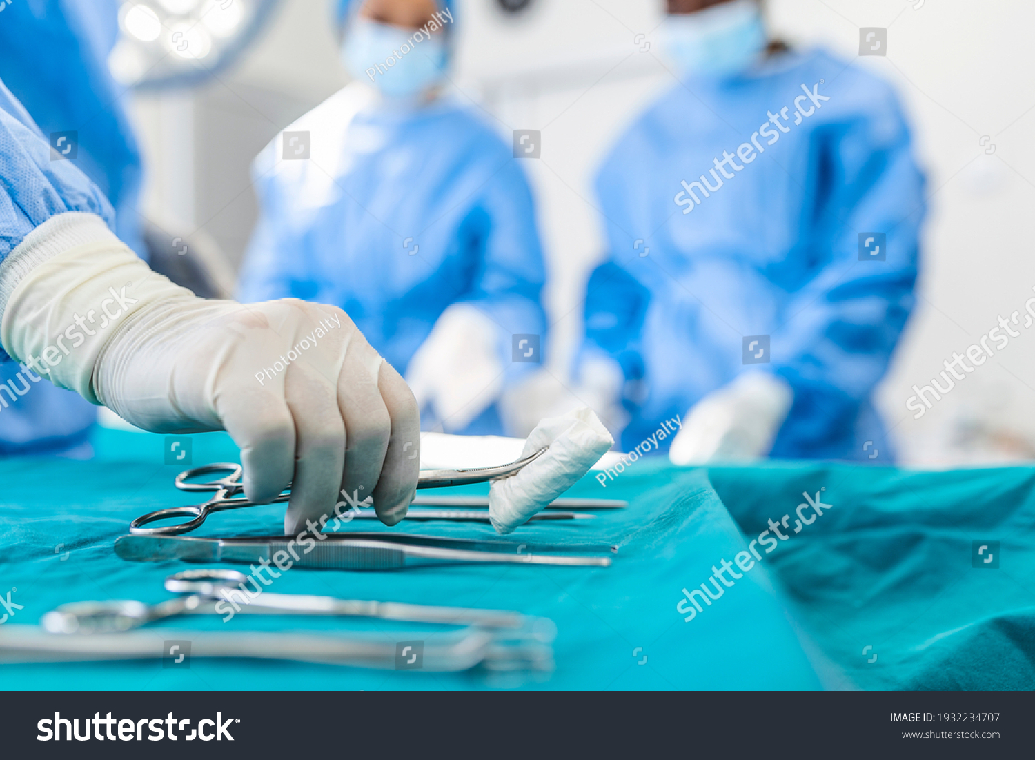 Nurse hand taking surgical instrument for group of surgeons at background operating patient in surgical theatre. Steel medical instruments ready to be used. Surgery and emergency concept #1932234707