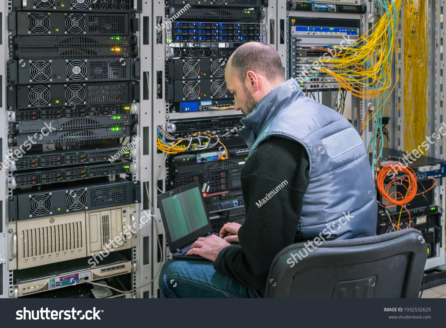 A man with a laptop sits in a server room. A technician works near the racks of a modern data center. The system administrator configures the computer hardware. #1932532625