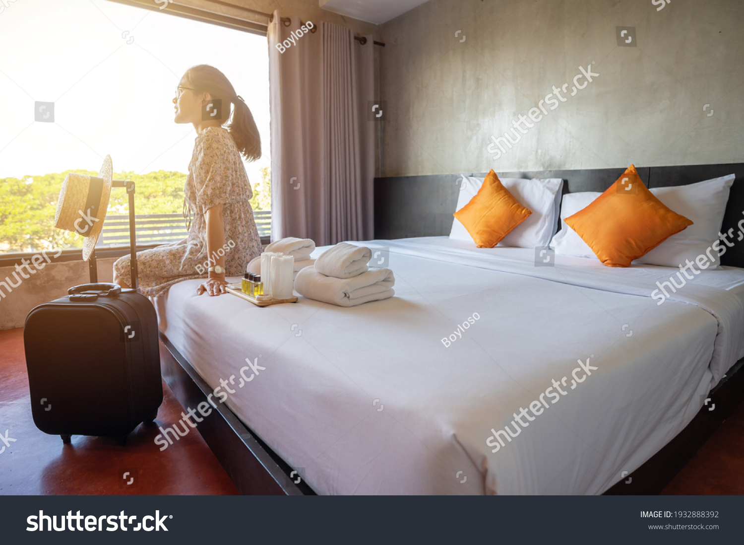 Tourist woman sitting on bed nearly window, looking to beautiful view with her luggage in hotel bedroom after check-in. Conceptual of travel and accommodation. #1932888392