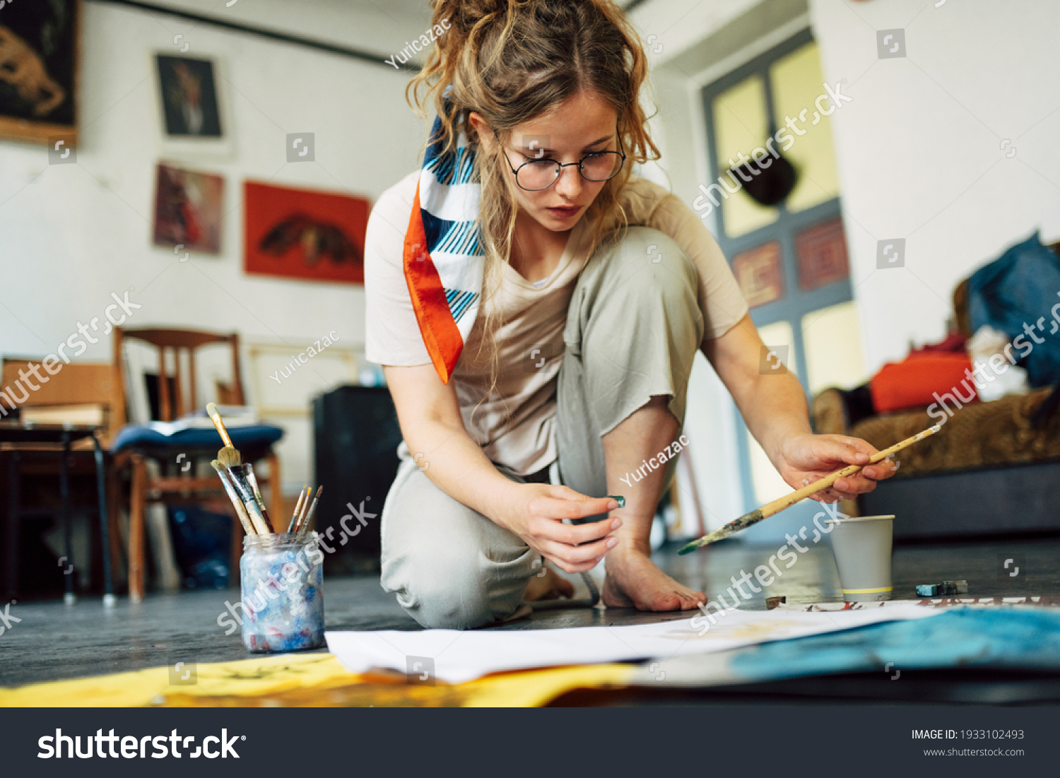 Horizontal image of a pretty female artist sitting on the floor in the art studio and painting on paper with a brush. A woman painter with glasses painting with watercolors in the workshop. #1933102493