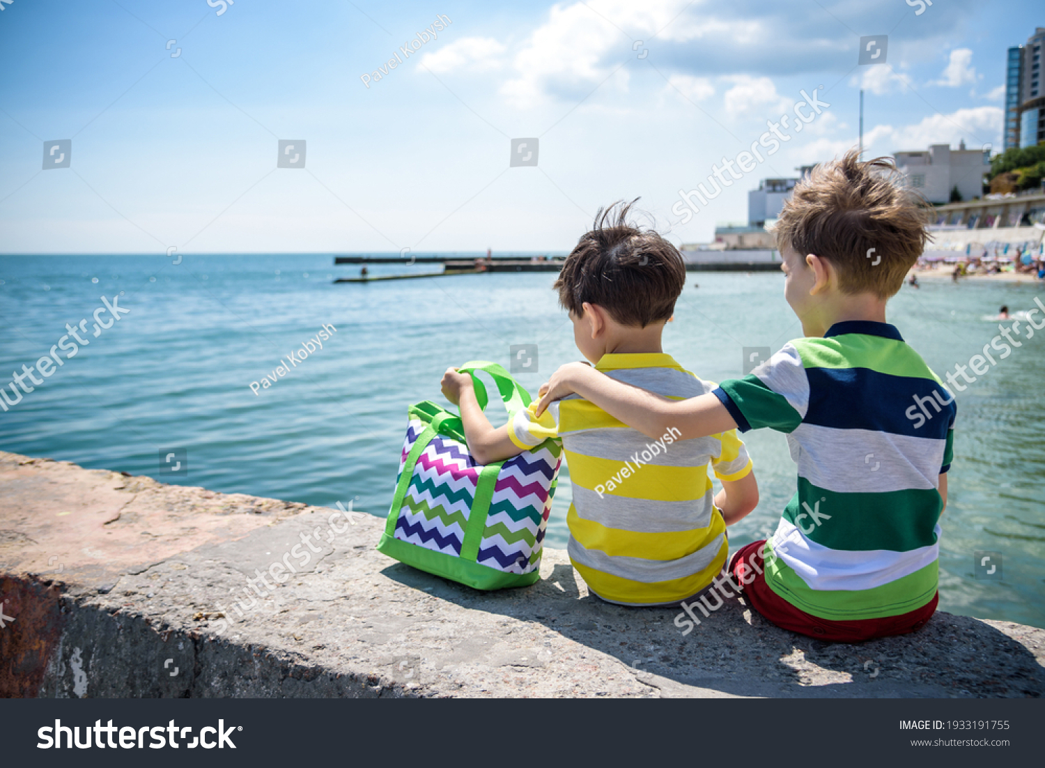 Two beautiful little boy sibling brothers in vest and shorts sitting on breakwater against the sea and shore in summer. Preschool children best friends enjoying summer vacations on sea. #1933191755