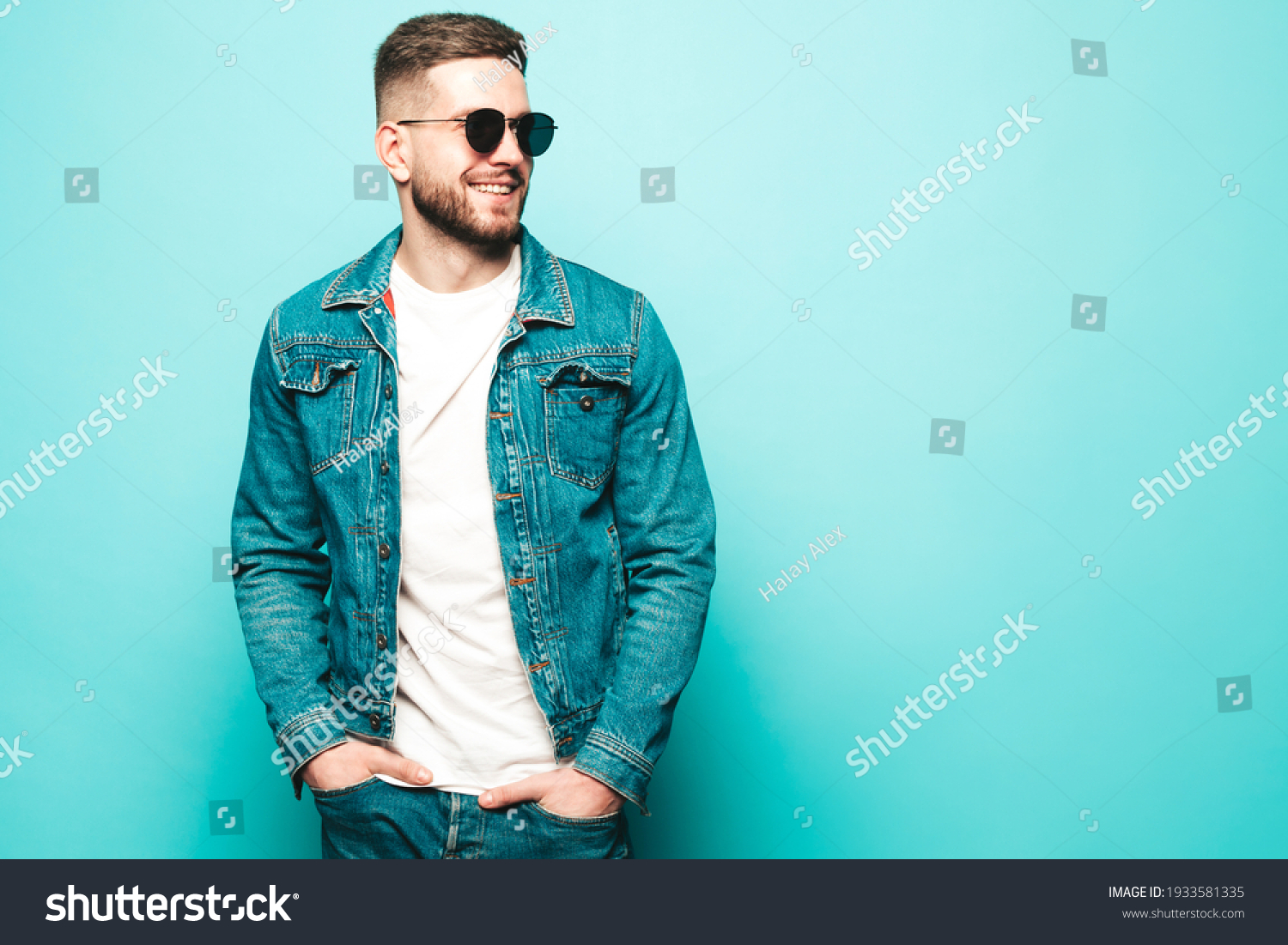 Portrait of handsome smiling stylish hipster lambersexual model.Man dressed in jacket and jeans. Fashion male posing near blue wall in studio in sunglasses #1933581335