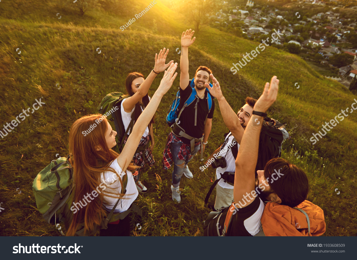 High angle of team of happy young tourists reaching out for high five while hiking on green grassy hill in the evening. From above of group of smiling people with backpacks having fun on trekking tour #1933608509