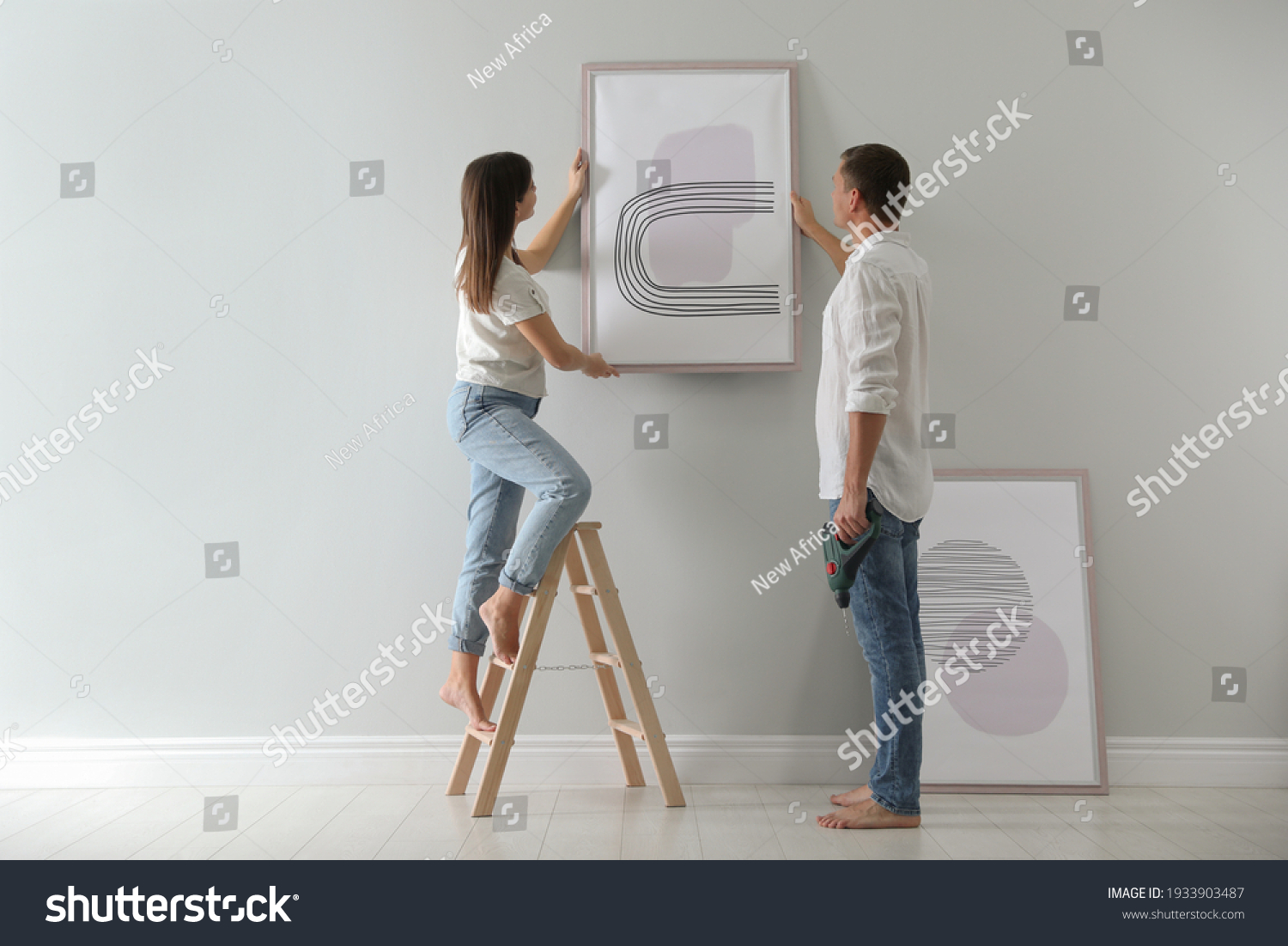Couple hanging picture on wall together in room. Interior design #1933903487