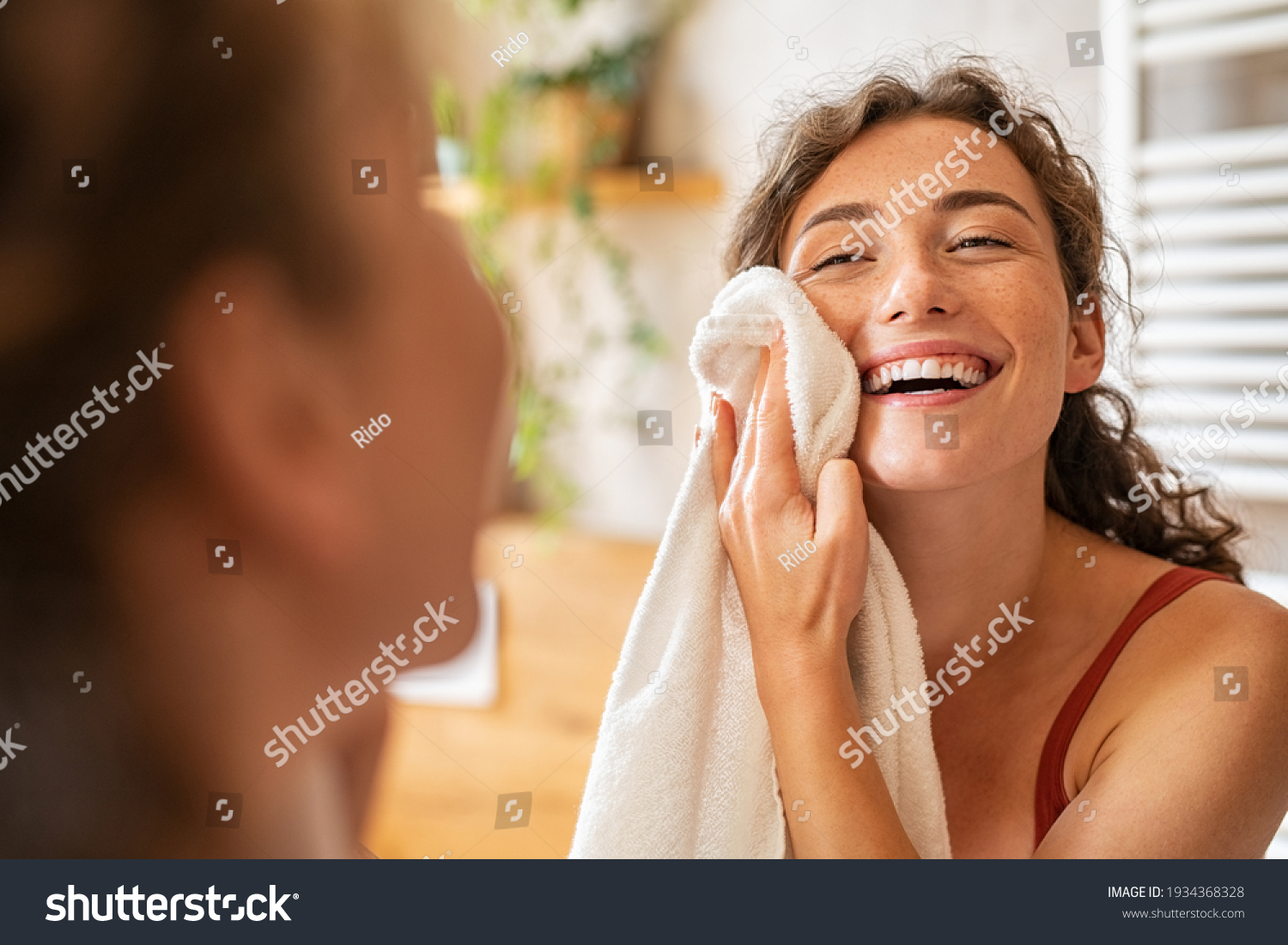 Young woman wiping her face with towel after waking up in the morning. Beautiful happy smiling girl holding towel near facial skin after washing face. Happy woman cleaning and drying skin with napkin. #1934368328