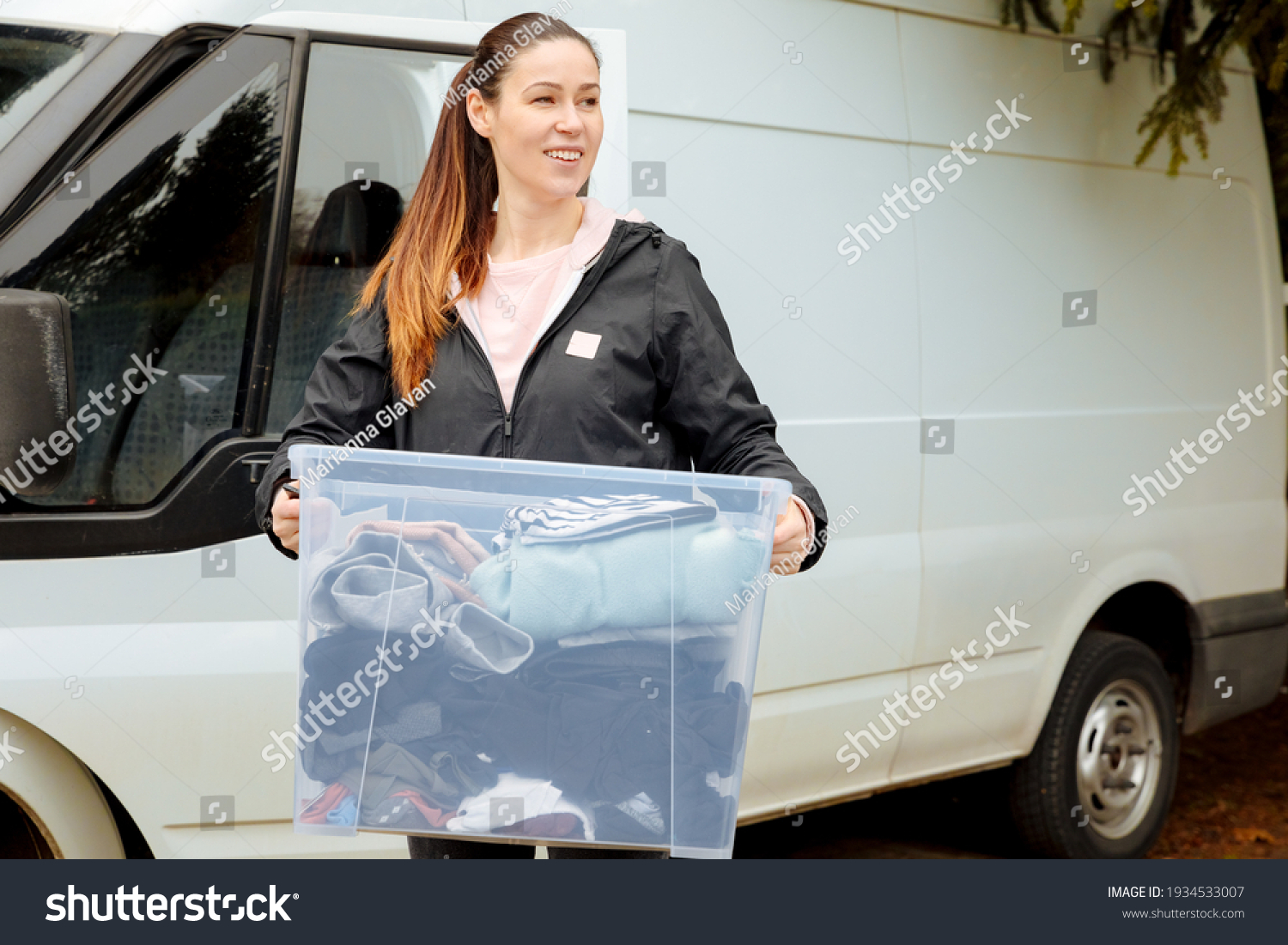 Young cute woman carries a plastic box with clothes. Volunteers provide clothing for refugees and the homeless. Deliver clean clothes with a delivery truck. Volunteer helps with the distribution  #1934533007