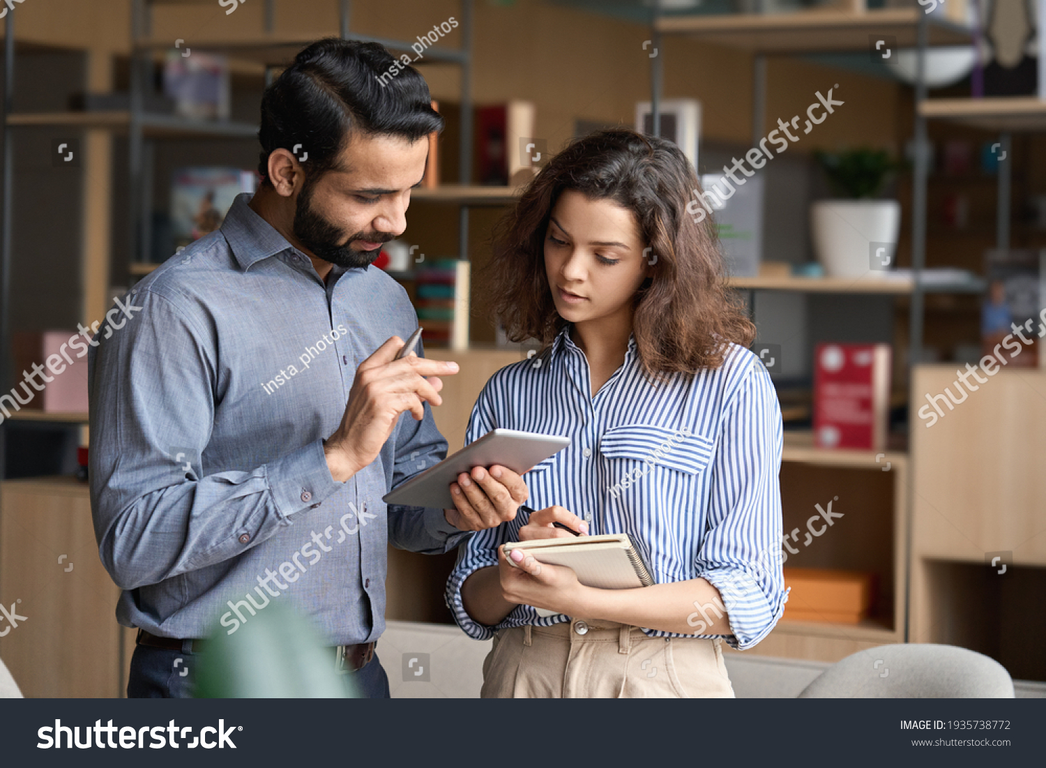 Indian manager holding digital tablet having discussion with latin employee meeting in office lobby. Two diverse professionals talking using pad discussing project strategy, checking corporate plan. #1935738772