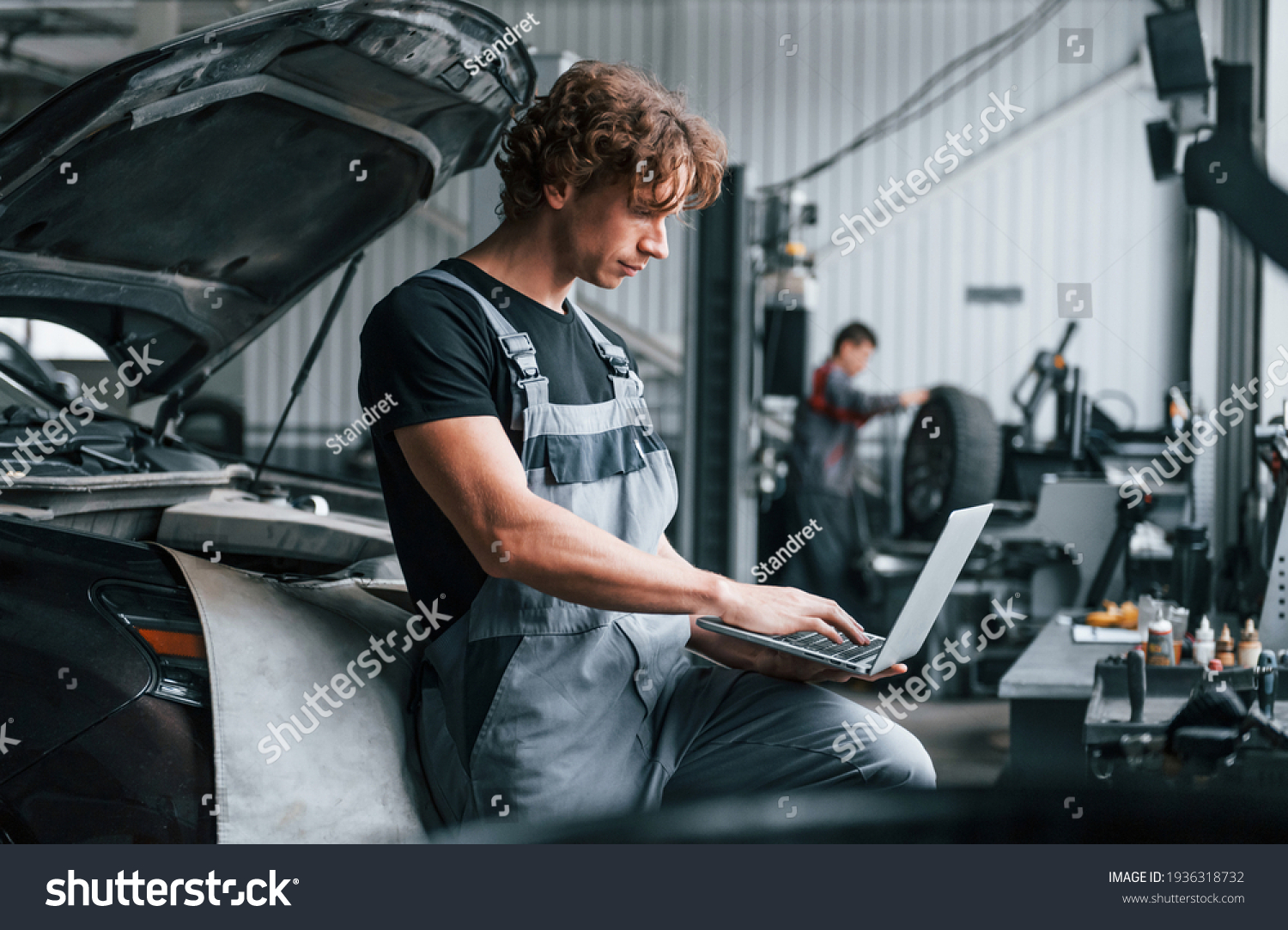 With laptop. Adult man in grey colored uniform works in the automobile salon. #1936318732