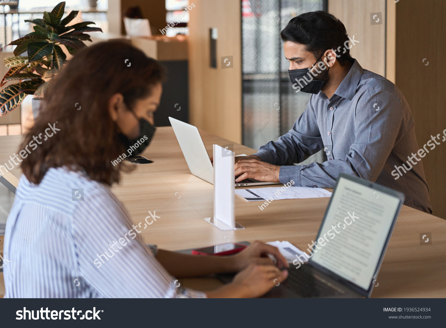 Indian business man student wearing face mask working on laptop safe distancing. Diverse people in facemasks using computers sitting at table with social distance sign in office coworking or cafeteria #1936524934