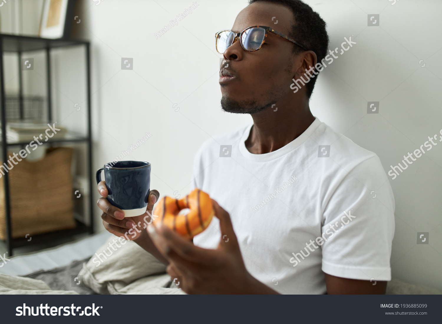 Indoor shot of stylish hungry young dark skinned man in glasses eating glazed orange doughnut with great relish, closing eyes with pleasure, enjoying every bite of sweet dessert, drinking morning tea #1936885099