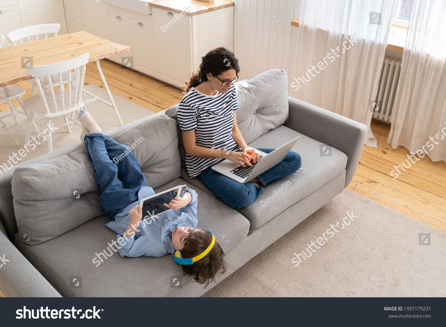Family, quarantine, distance job concept. Happy mother freelancer remote work from home office on laptop during lockdown, sitting on sofa, little child playing on digital tablet and wear headphones.  #1937179231