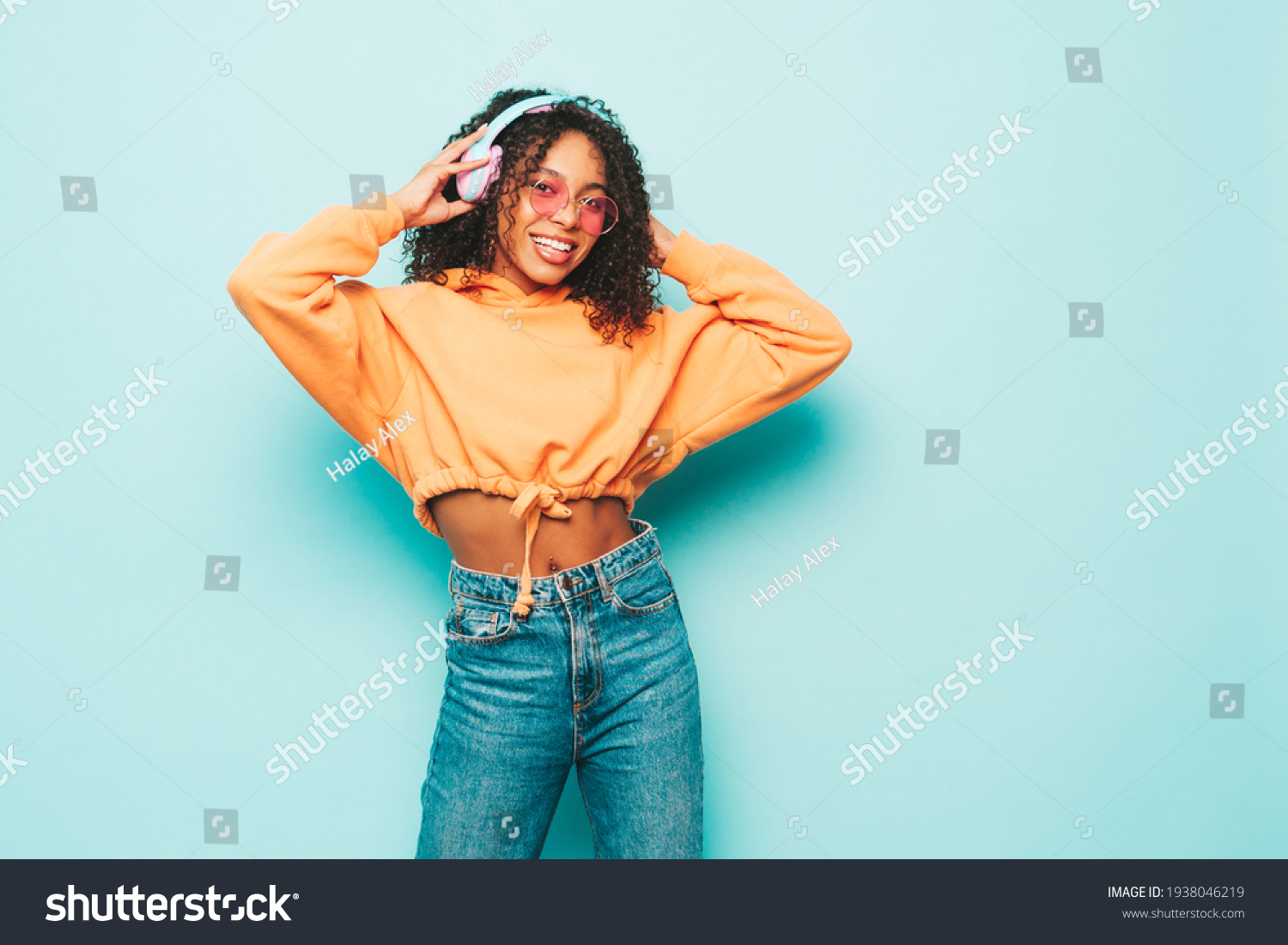 Beautiful black woman with afro curls hairstyle.Smiling model in orange hoodie and jeans.Sexy carefree female listening music in wireless headphones.Posing in studio near blue wall #1938046219