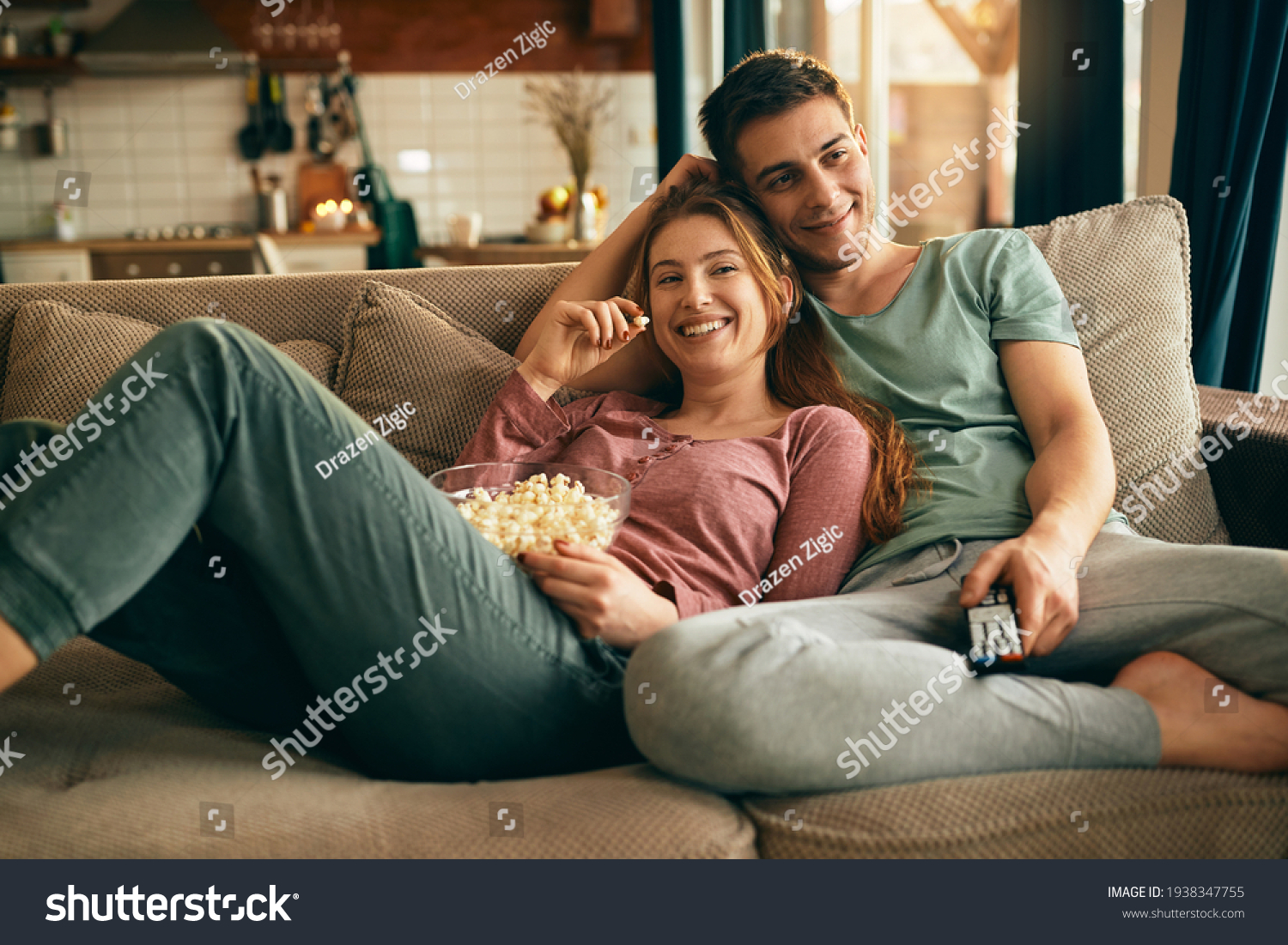 Young happy couple watching movie on TV and eating popcorn while relaxing in the living room.  #1938347755