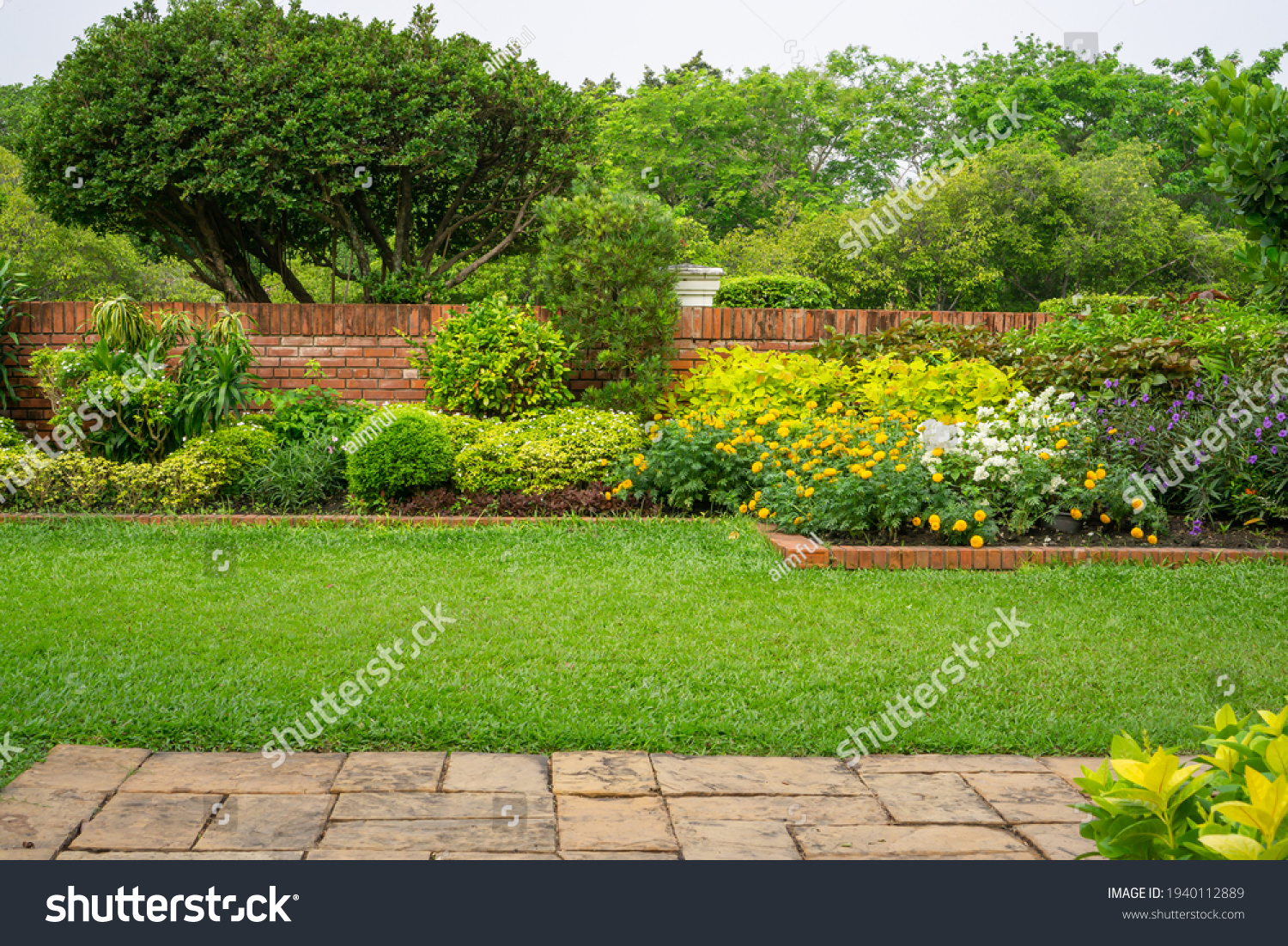 Backyard English cottage garden, colorful flowering plant and green grass lawn, brown pavement and orange brick wall, evergreen trees on background, in good care maintenance landscaping in park  #1940112889