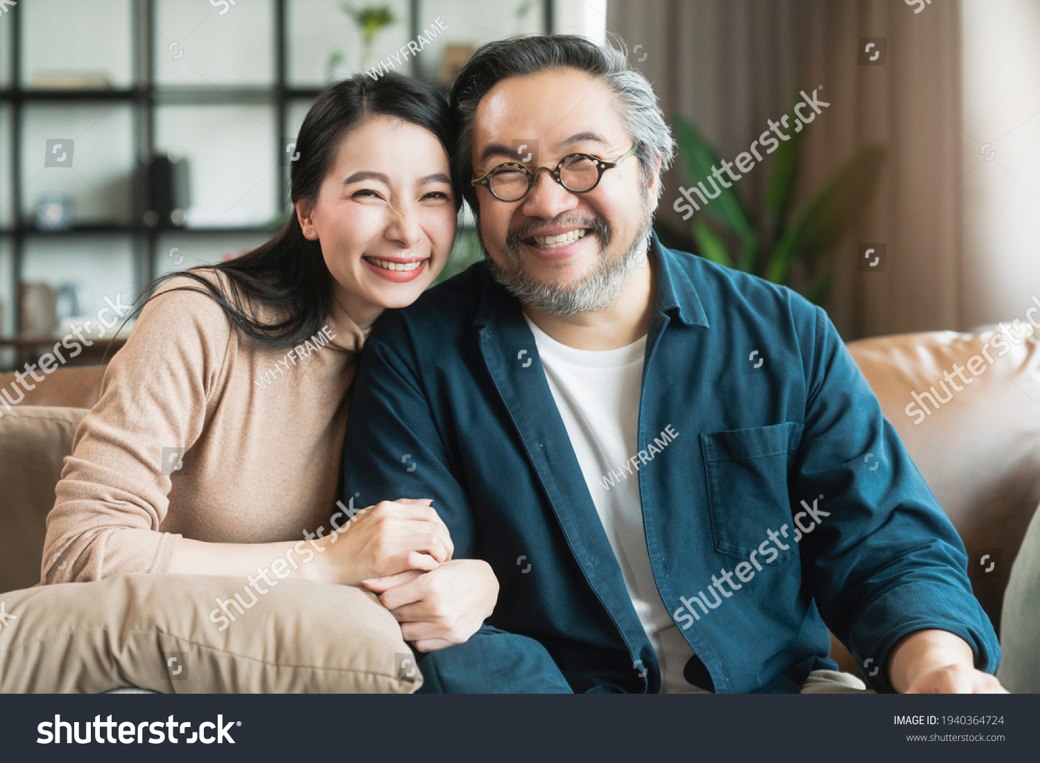Portrait of Asian mature couple sitting and smiling in living room. wife woman hand hold husband arm from behind and look at camera with happiness and cheerful safty amd insurance family concept #1940364724
