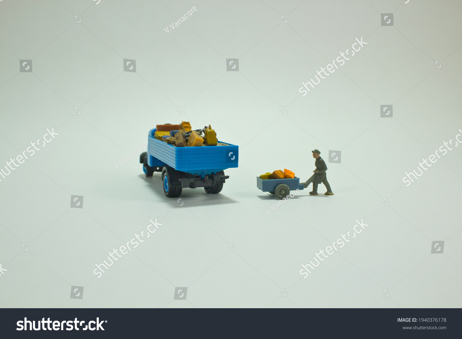 View of a miniature figure scene. A blue truck stands fully loaded with luggage while a worker pushes a handcart with other luggage to the truck. White background. Loading and transportation concept #1940376178
