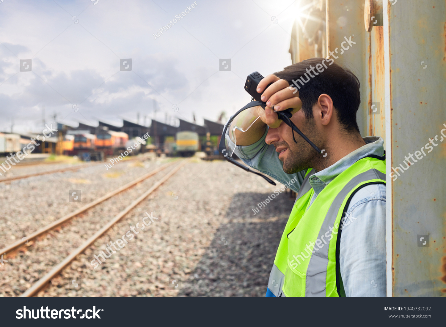 A Railway engineer or Rail transport technician wearing a green safety vest is standing to rest or working outdoors beside a freight train on a hot and sunny day. #1940732092
