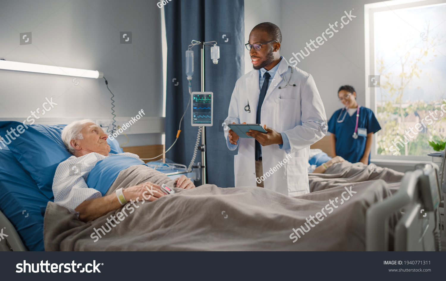 Hospital Ward: Friendly Black Doctor Talks with Elderly Caucasian Patient Resting in Bed, Asks Health Care Questions. Doctor Uses Tablet Computer. Old Man Fully Recovering after Successful Surgery #1940771311