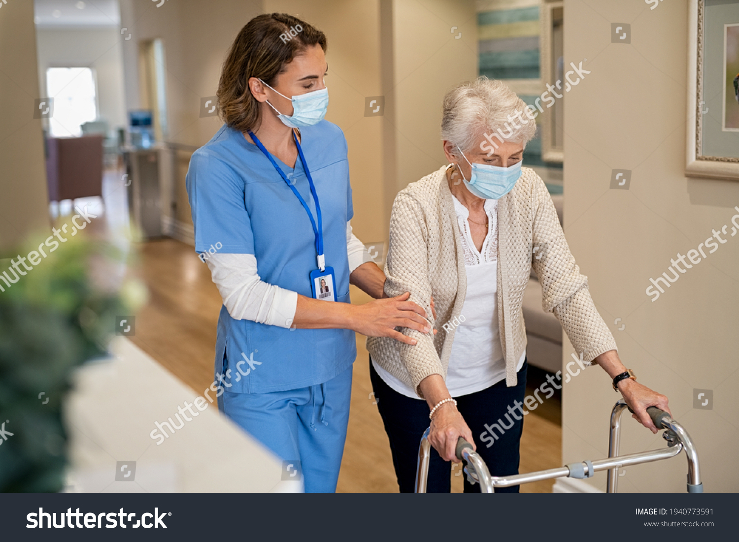 Smiling nurse with face mask helping senior woman to walk around the nursing home with walker. Young lovely nurse helping old woman with surgical mask for safety against covid-19 using a walking frame #1940773591
