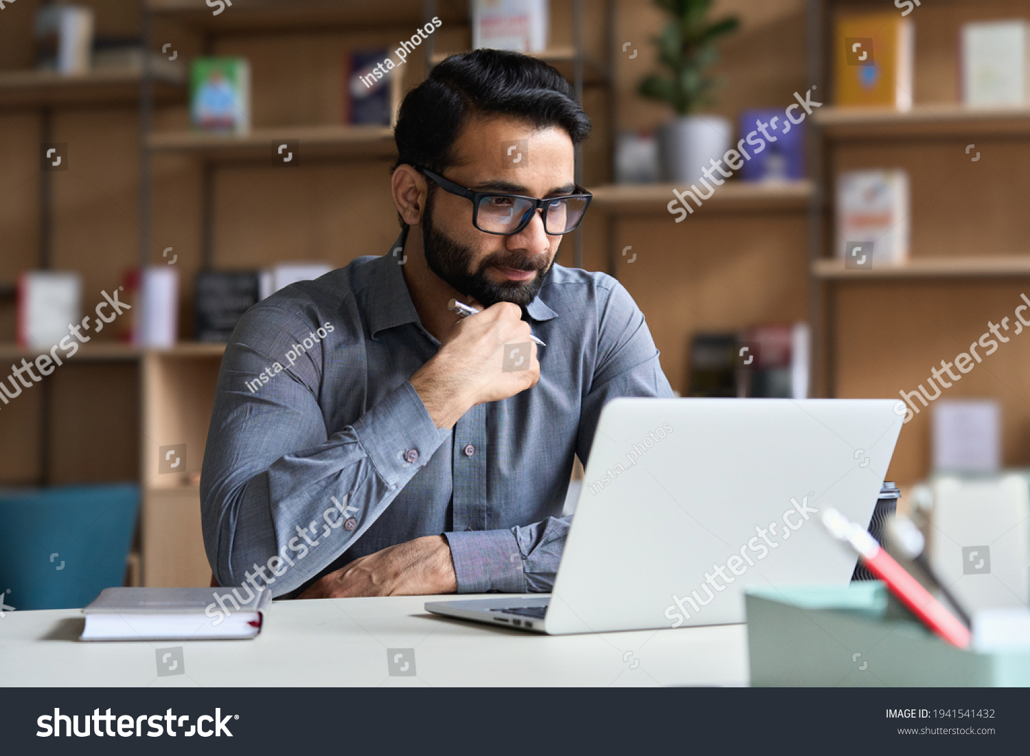 Young serious indian professional business man, focused ethnic male student wearing glasses working on laptop, remote studying using computer looking at screen watching seminar webinar at home office. #1941541432