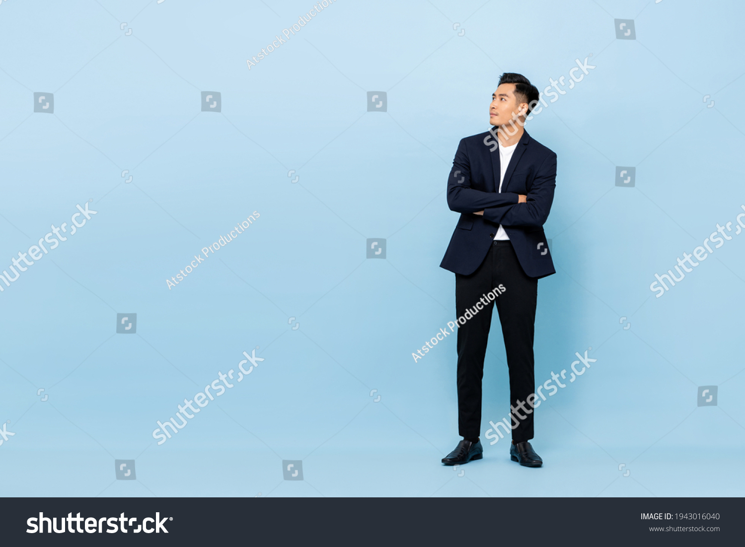 Full length portrait of young handsome southeast Asian businessman with arms crossed looking up sideway to copy space on light blue studio background #1943016040