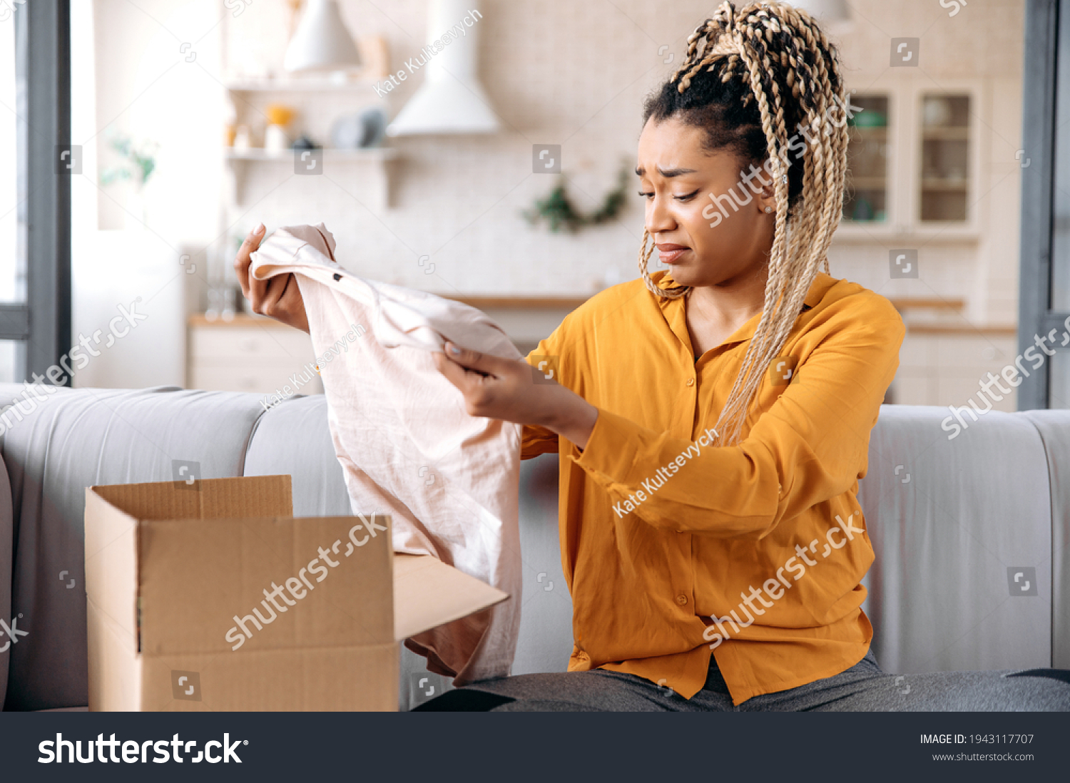 A confused sad african american girl unpacking her parcel at home. Surprised young woman got an unexpected order, girl is upset with her parcel, unsuccessful online shopping. Home delivery #1943117707