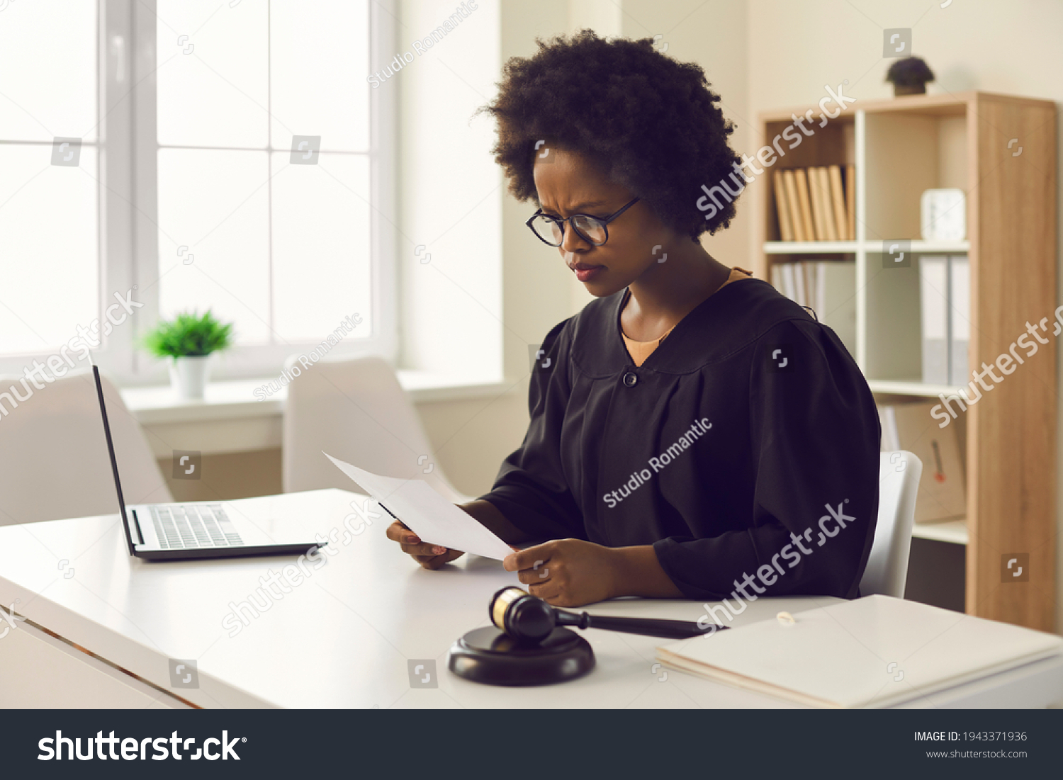 Frowning young african american female judge reading paper document sitting at desk front of laptop in courtroom. Legal trial or tribunal, paperwork and criminal data or jury order investigation #1943371936