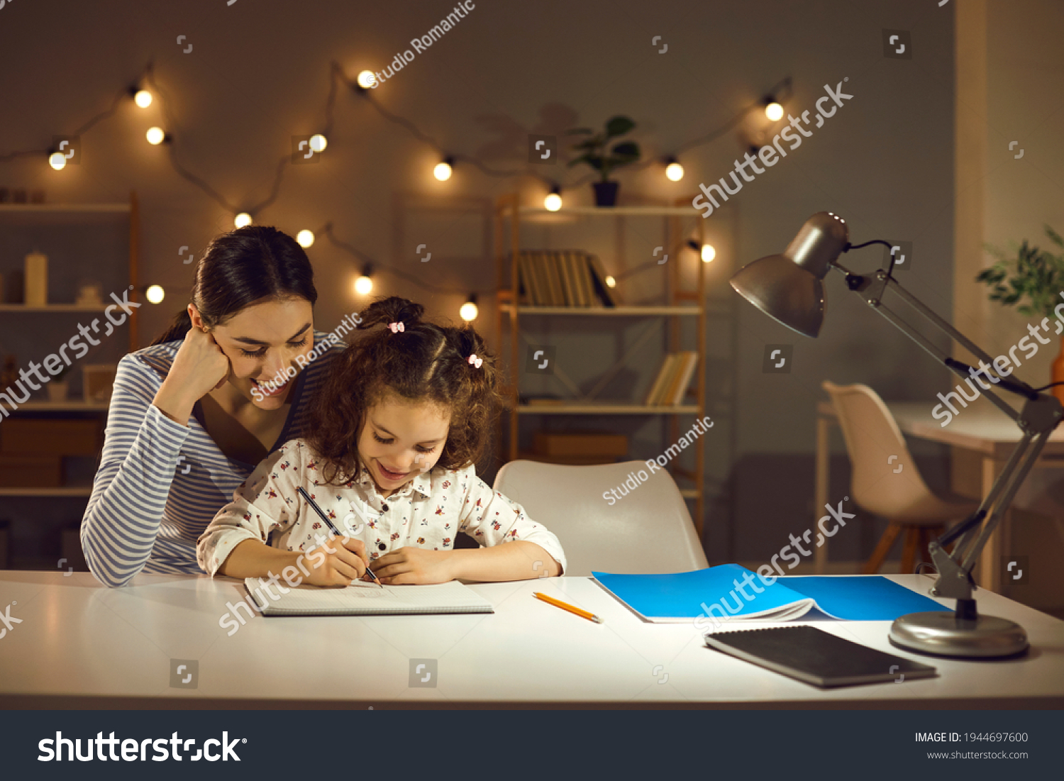 Parent helping child. Happy family doing homework in the evening. Mother and daughter working on school assignment sitting at desk with lamp in cozy dark room with LED lights. Kids learning concept #1944697600