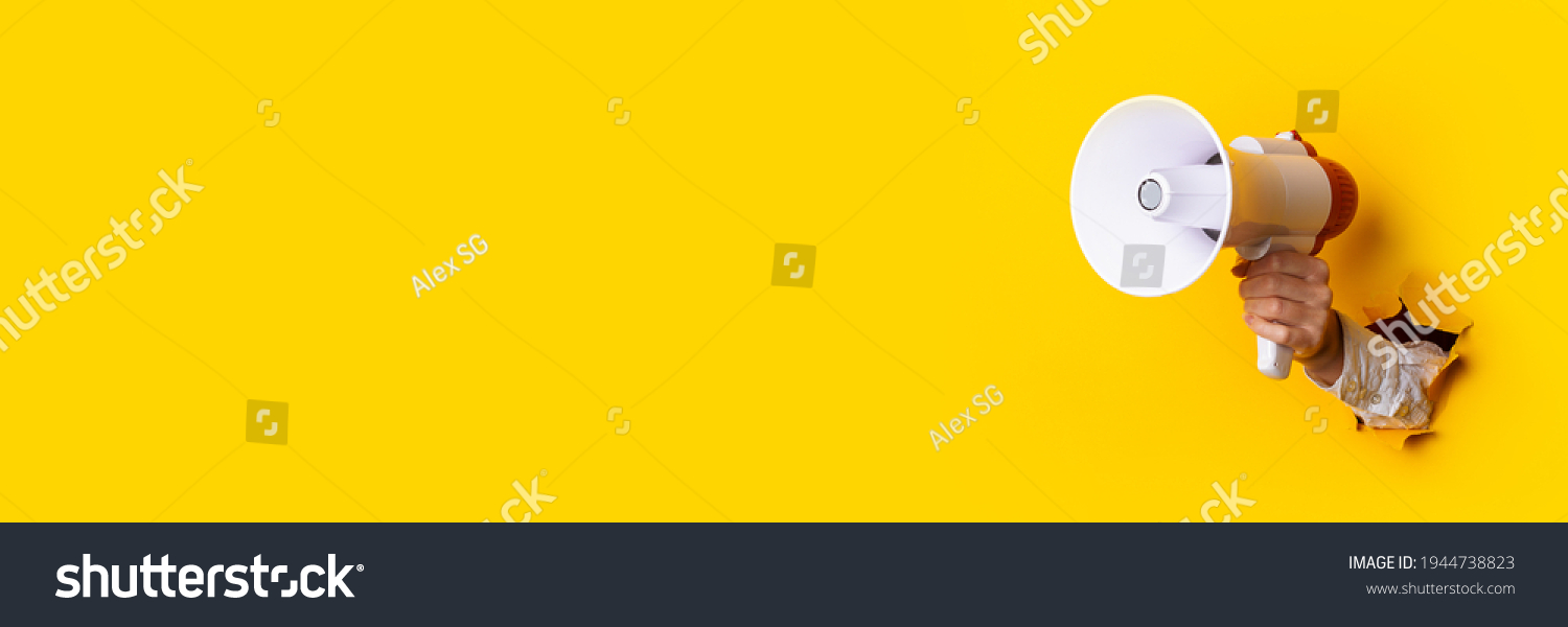 Hand holds a megaphone from a hole in the wall on a yellow background. Concept of hiring, advertising something. Banner. #1944738823