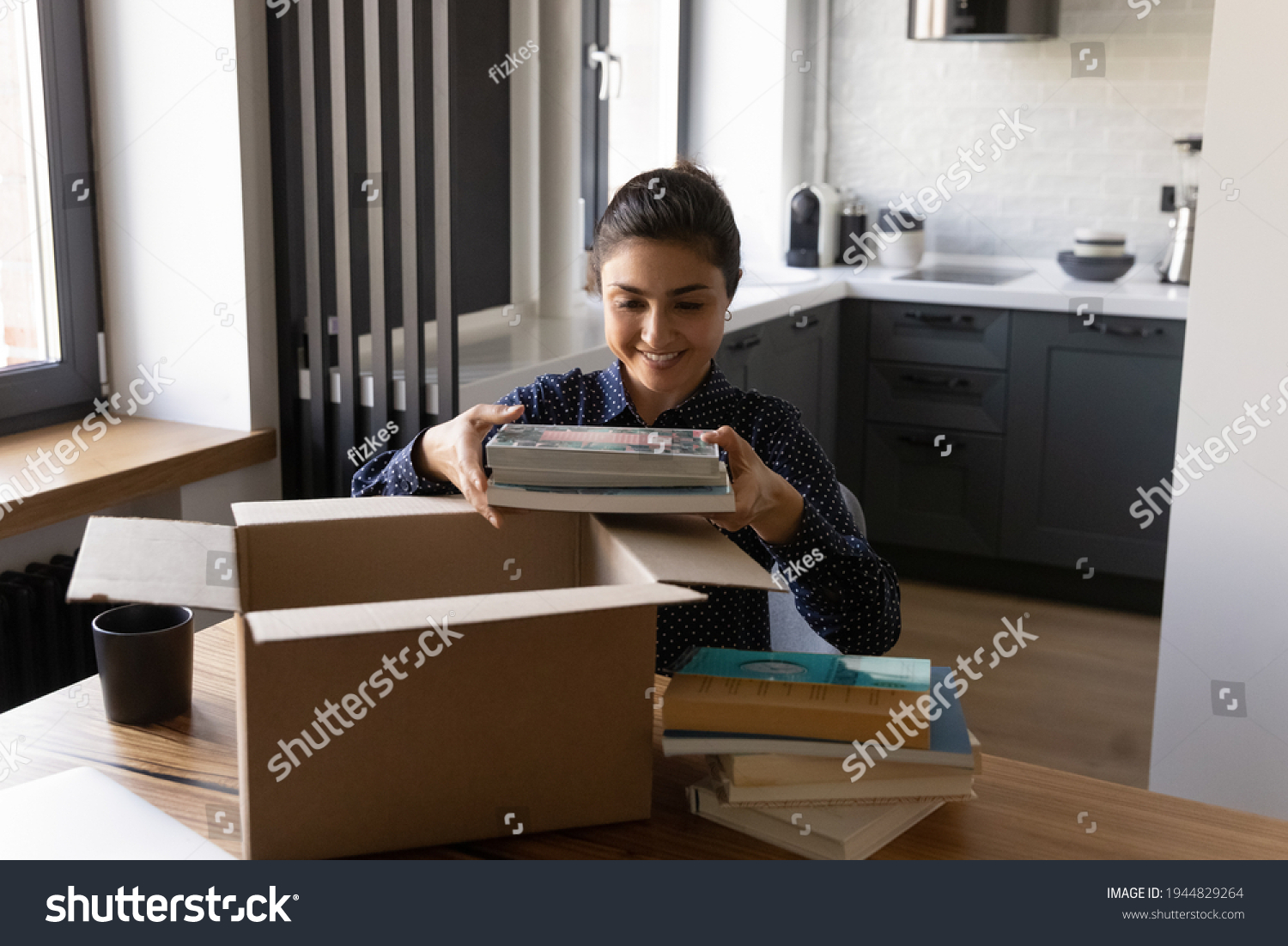 Smiling Indian woman unpacking parcel, holding books, sitting at table, satisfied customer received online store order, delivery service, happy female packing belongings, preparing for relocation #1944829264