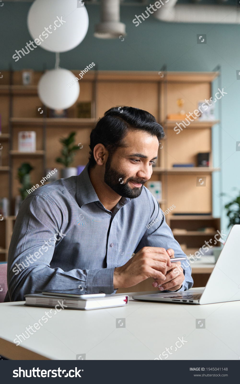 Smiling bearded indian businessman working on laptop at home office. Young indian student using computer remote studying, virtual training on video call meeting, watching online webinar or seminar. #1945041148