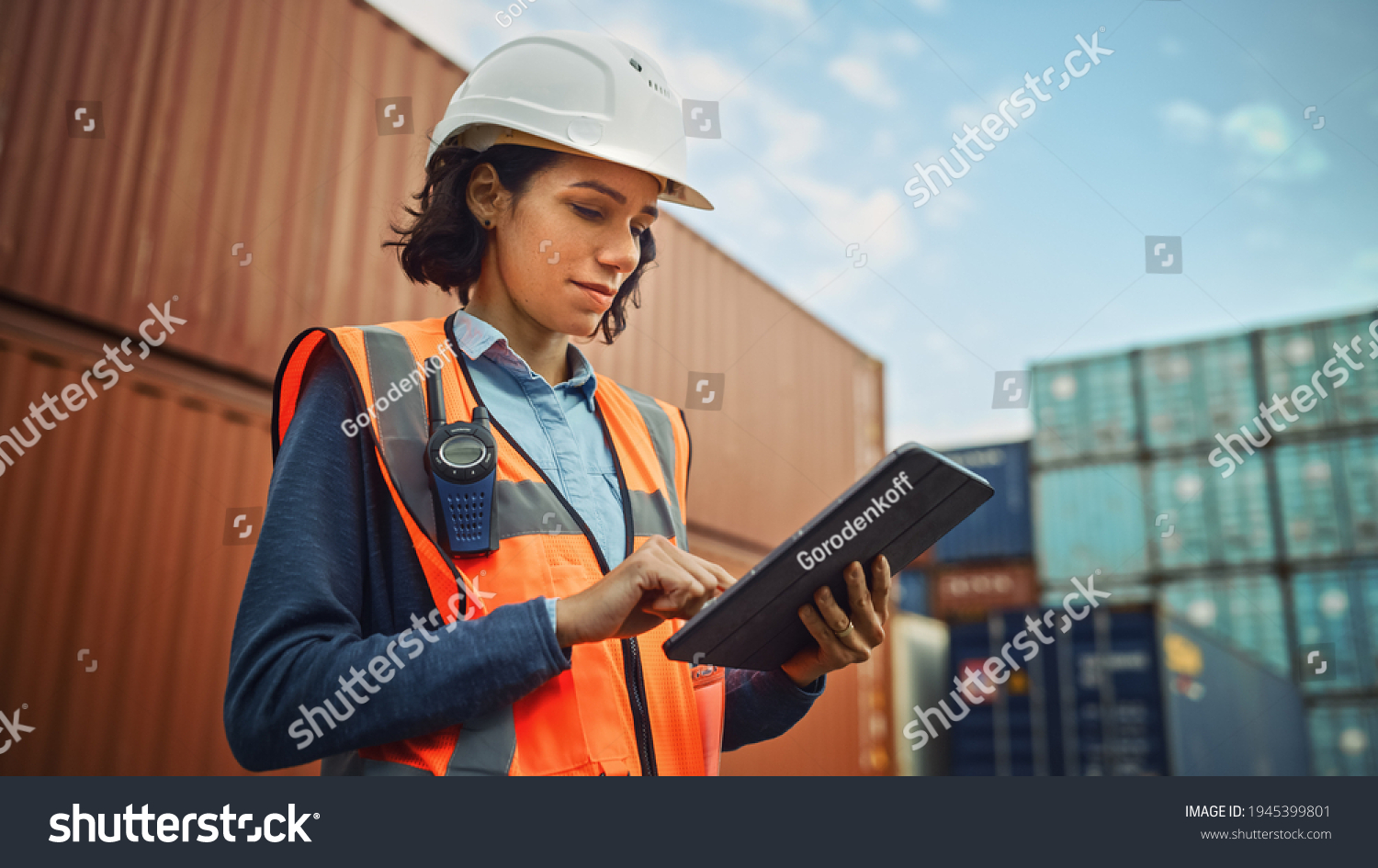 Smiling Portrait of a Beautiful Latin Female Industrial Engineer in White Hard Hat, High-Visibility Vest Working on Tablet Computer. Inspector or Safety Supervisor in Container Terminal. #1945399801