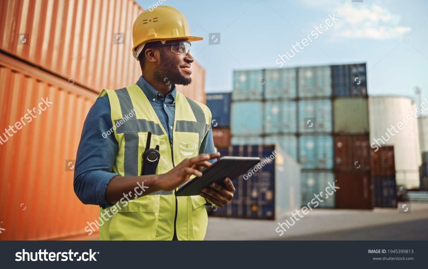 Smiling Portrait of a Handsome African American Black Industrial Engineer in Yellow Hard Hat and Safety Vest Working on Tablet Computer. Foreman or Supervisor in Container Terminal. #1945399813
