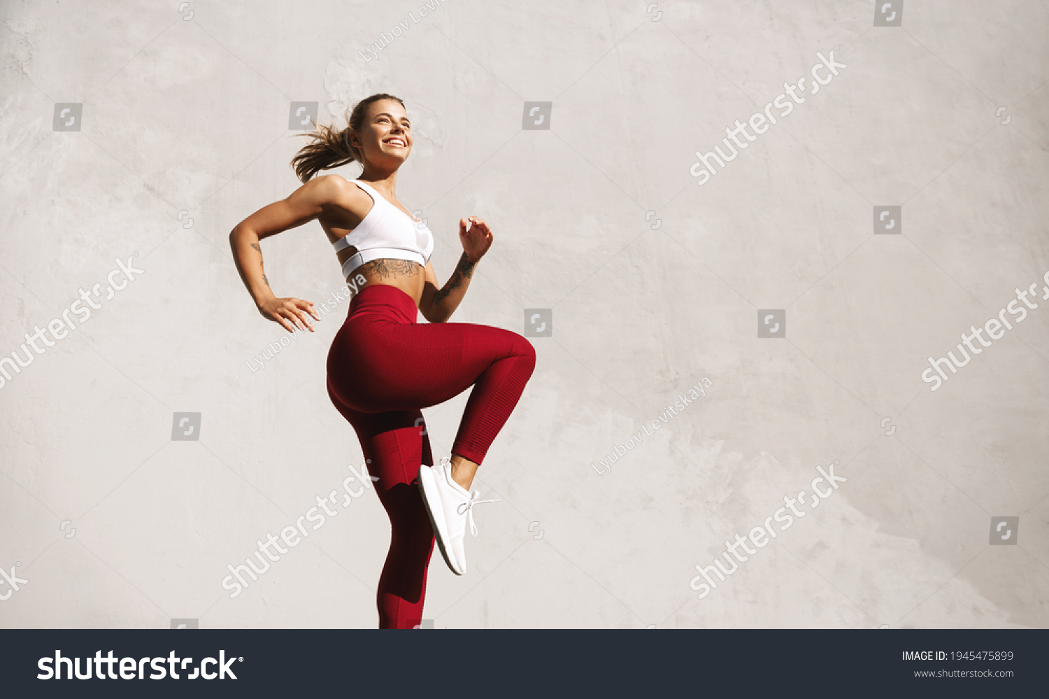 Fit woman exercising outdoors. Healthy young female athlete doing fitness workout. Sportswoman raising leg, do functional training outside on bright sunny day, smiling pleased, wearing sport outfit #1945475899
