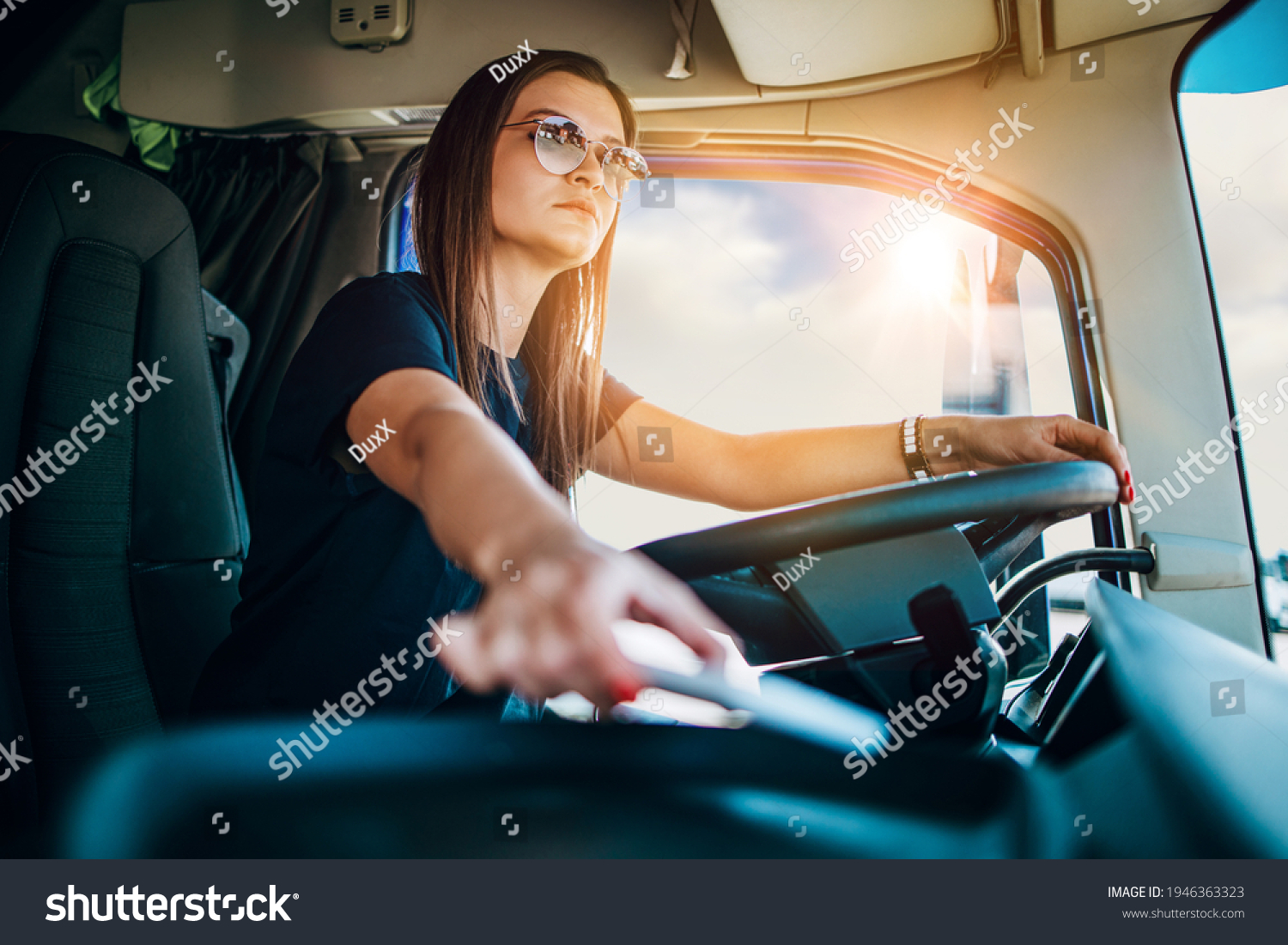 Portrait of beautiful young woman professional truck driver sitting and driving big truck. She is dangerously trying to take smart phone while driving. #1946363323