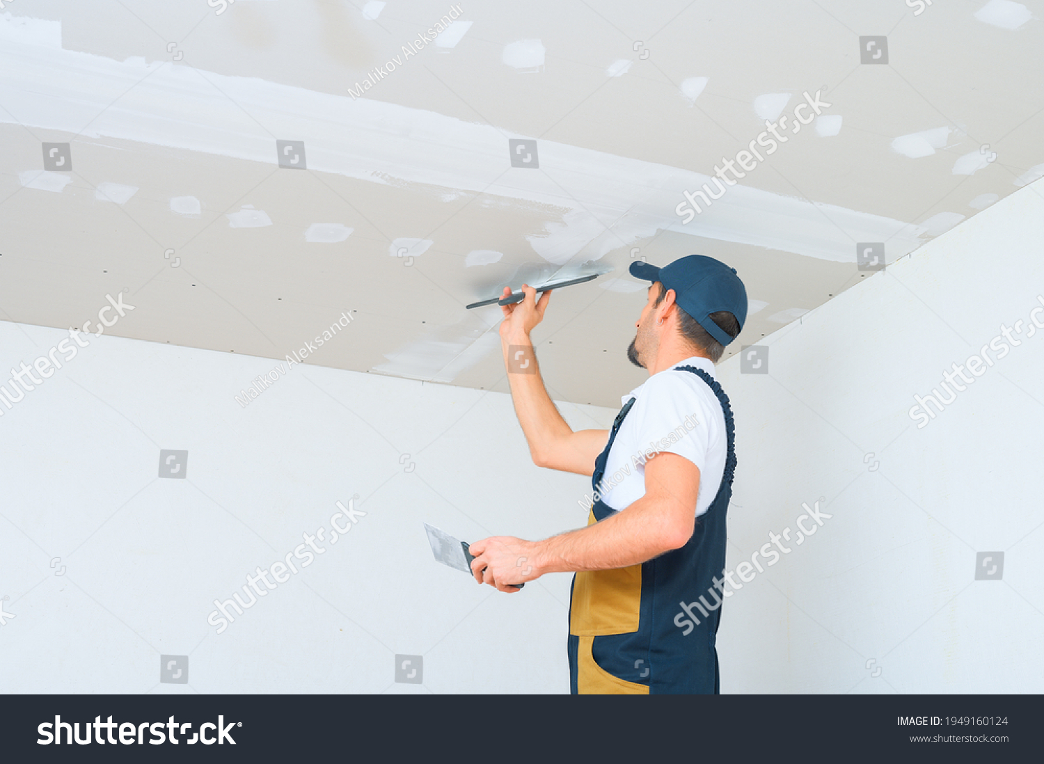 A uniformed worker applies putty to the drywall ceiling. Putty of joints of drywall sheets. #1949160124