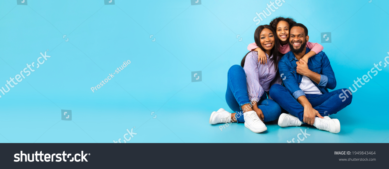 Happy Loving Family. Black man, woman and girl sitting on the floor isolated on blue studio wall. Smiling daughter emracing her mum and dad from behind, posing at camera, banner, panorama, copy space #1949843464
