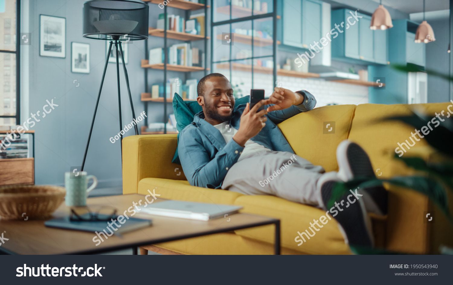 Excited Black African American Man Using Smartphone while Resting on a Sofa in Living Room. Happy Man Smiling at Home and Chatting to Colleagues and Clients Over the Internet. Using Social Networks. #1950543940