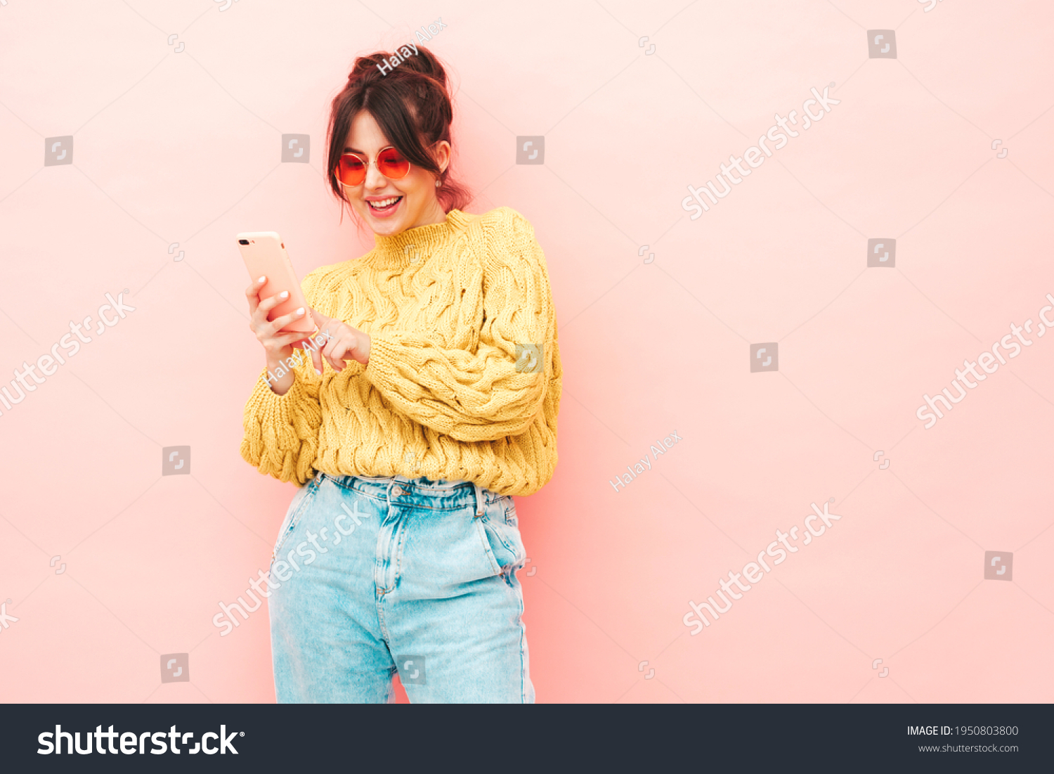 Young beautiful smiling female in trendy summer yellow hipster sweater and jeans.Sexy carefree woman posing near pink wall in studio.Positive model having fun. Looking at smartphone screen, using app #1950803800
