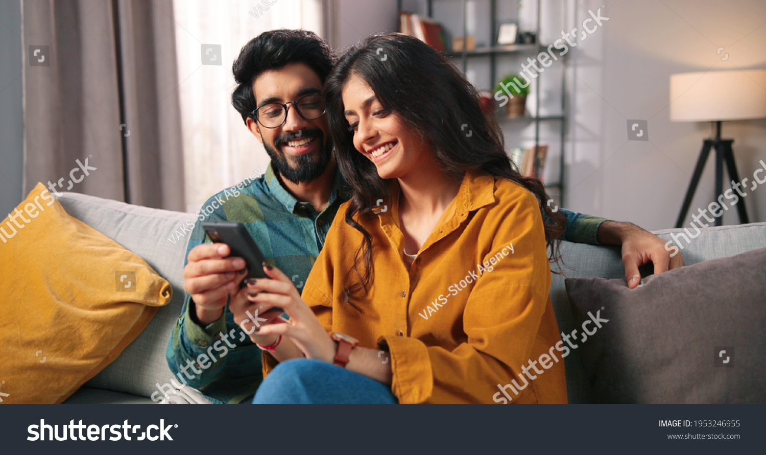 Portrait of cheerful positive young lovely couple smiling spending time together at home sitting on sofa typing on smartphone, searching internet using social network app on cellphone, family concept #1953246955