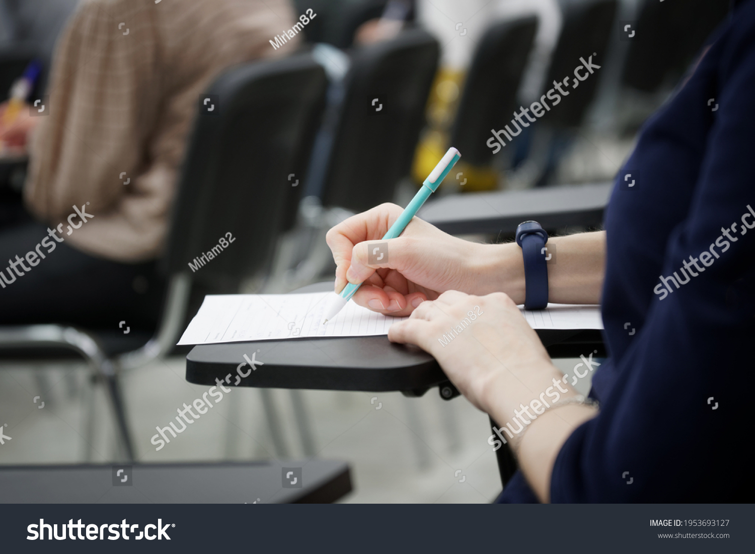 A girl writes a dictation or fills out documents in the audience, sitting on a school chair with a writing stand. Close-up. No face #1953693127