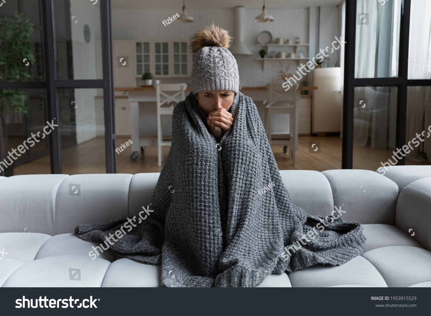 Frozen. Sad latina female sit on couch at freezing cooled studio flat in warm cap and blanket shiver tremble with cold. Unhappy young lady spend time at home feel bad suffer of heating system problems #1953915529